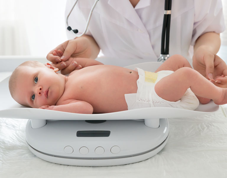 Um bebê deitado na balança tem seu peso medido, enquanto uma médica segura sua mãozinha e o mantém calmo.