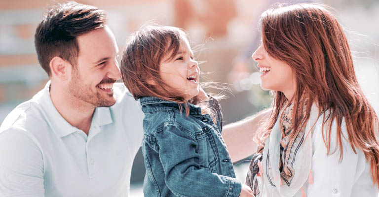 A imagem mostra uma família sorridente. O pai, à esquerda, tem cabelo curto castanho e barba rala. A filha, no centro, tem cabelo comprido castanho e olha para sua mãe, à direita, que também tem cabelo comprido castanho.