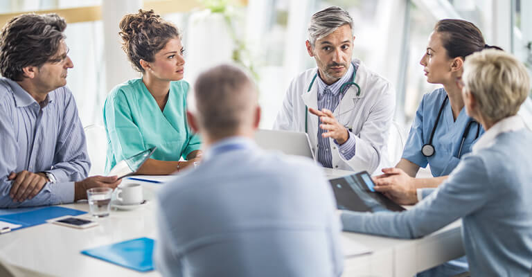 Seis médicos reunidos ao redor de uma mesa conversam. Em foco, há duas médicas de cabelos presos, um médico de óculos e camisa listrada azul e branca, e, no centro, um outro médico de cabelo e barba grisalhos, a quem todos escutam.