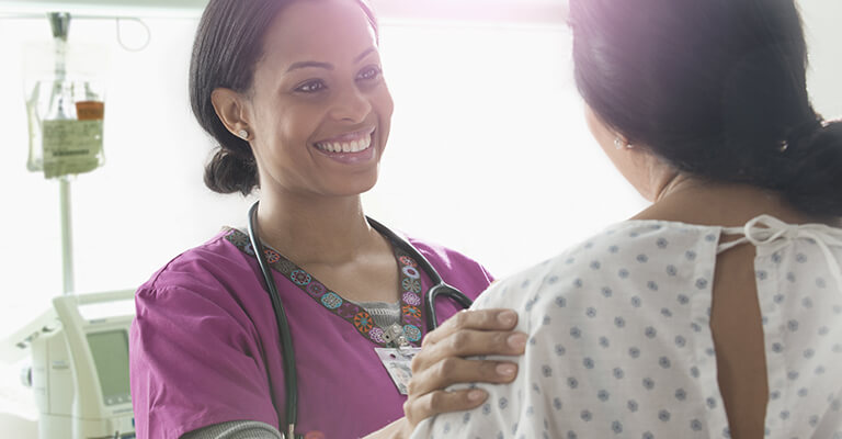 A imagem mostra uma médica de cabelo preso, sorrindo para uma paciente e demonstrando apoio através de um leve toque no ombro.